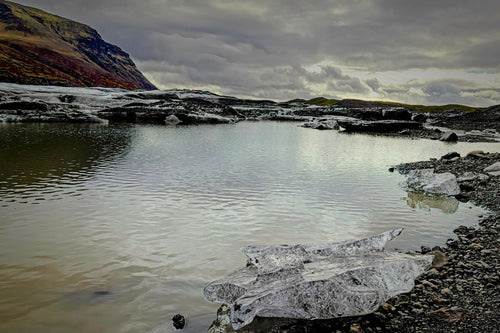 Chunks of glacial ice stranded on a black gravel shore of a glacial lagoon, a mountain and glacier tongue reflected in the still water under an overcast sky