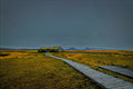 A wooden boardwalk leading across golden grassland to a turf-roofed Icelandic farmhouse, distant mountains under a moody dusk sky