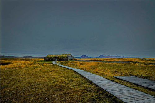 A wooden boardwalk leading across golden grassland to a turf-roofed Icelandic farmhouse, distant mountains under a moody dusk sky