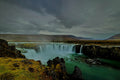 A wide horseshoe waterfall spilling from dark basalt cliffs into a turquoise river basin under brooding storm clouds