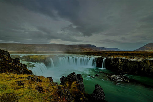 A wide horseshoe waterfall spilling from dark basalt cliffs into a turquoise river basin under brooding storm clouds