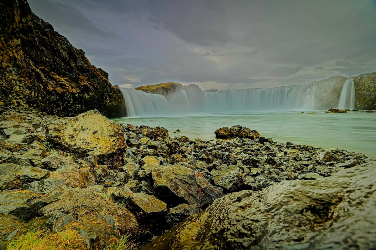 A curved waterfall softened to a veil by long exposure, seen low across jagged black basalt boulders under a moody grey sky