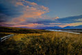 A wide Icelandic valley of windswept golden grass and a winding river, distant mountains under dramatic pink and blue sunset clouds