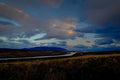 A braided river winding through a dark flat valley at dusk, pink-tinged storm clouds gathering over a distant mountain silhouette