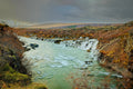 A row of small waterfalls feeding a turquoise river between autumn-colored lava-field banks, moody grey sky over open highland beyond