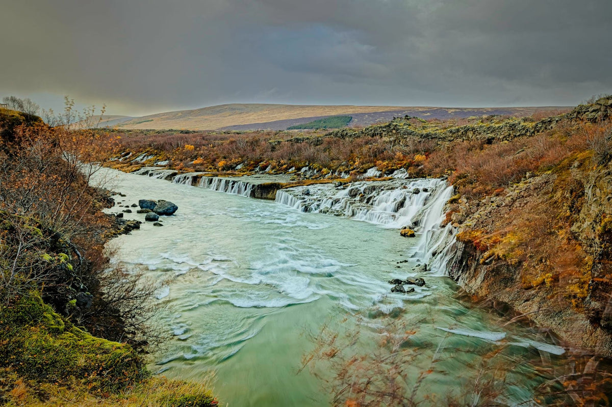 A row of small waterfalls feeding a turquoise river between autumn-colored lava-field banks, moody grey sky over open highland beyond
