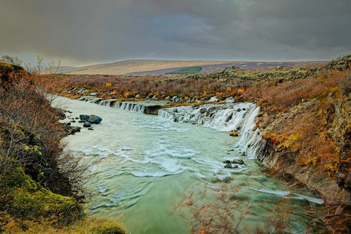 A row of small waterfalls feeding a turquoise river between autumn-colored lava-field banks, moody grey sky over open highland beyond