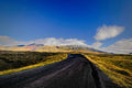 A dark road curving through golden highland moss toward a snow-dusted volcanic ridge under a deep blue sky