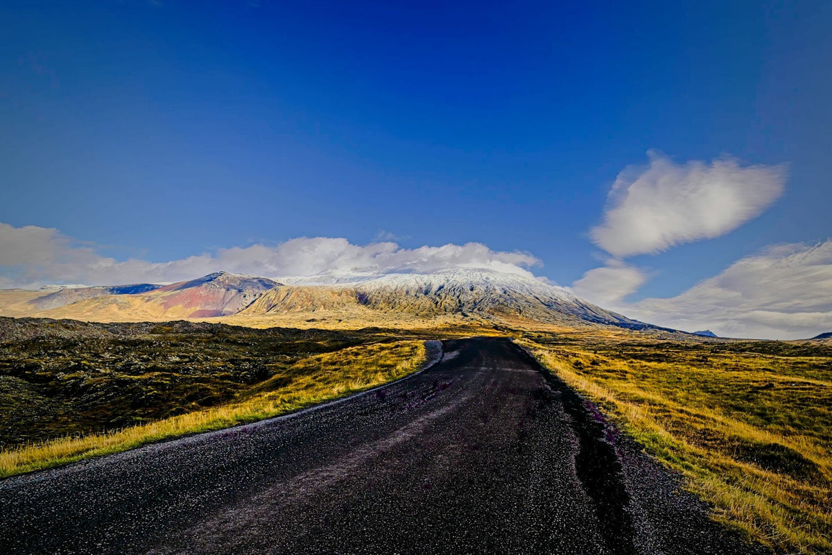 A dark road curving through golden highland moss toward a snow-dusted volcanic ridge under a deep blue sky