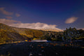 A snow-capped mountain wrapped in cloud beyond a black lava field scattered with rust-colored stones and a small still pool, deep blue evening sky