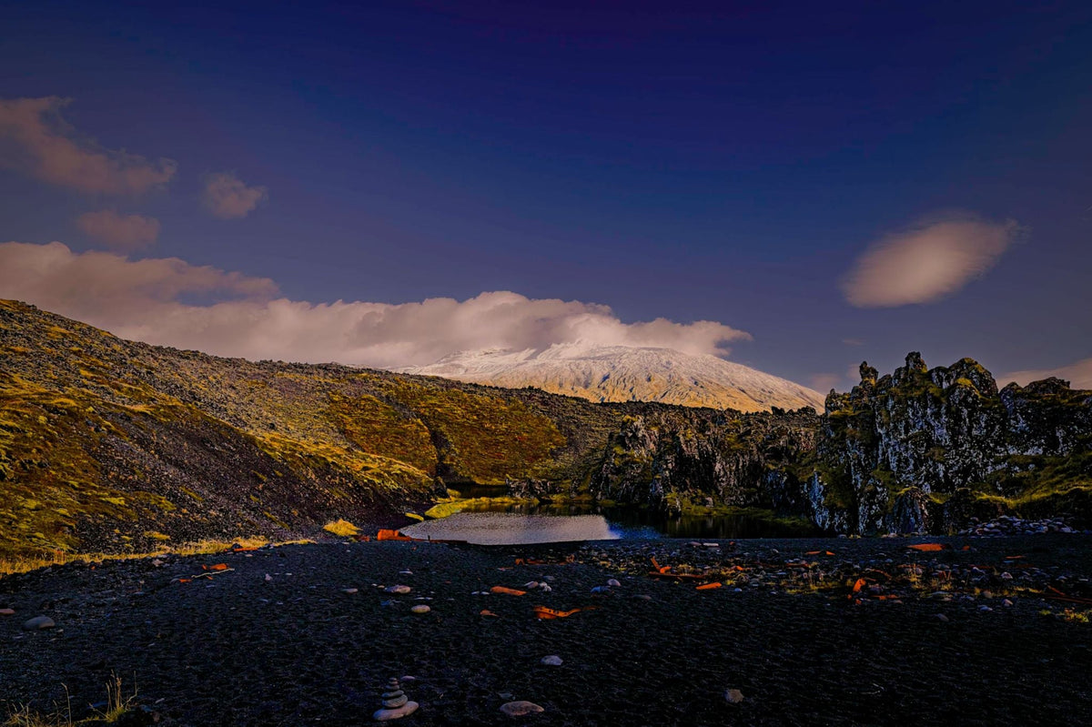 A snow-capped mountain wrapped in cloud beyond a black lava field scattered with rust-colored stones and a small still pool, deep blue evening sky
