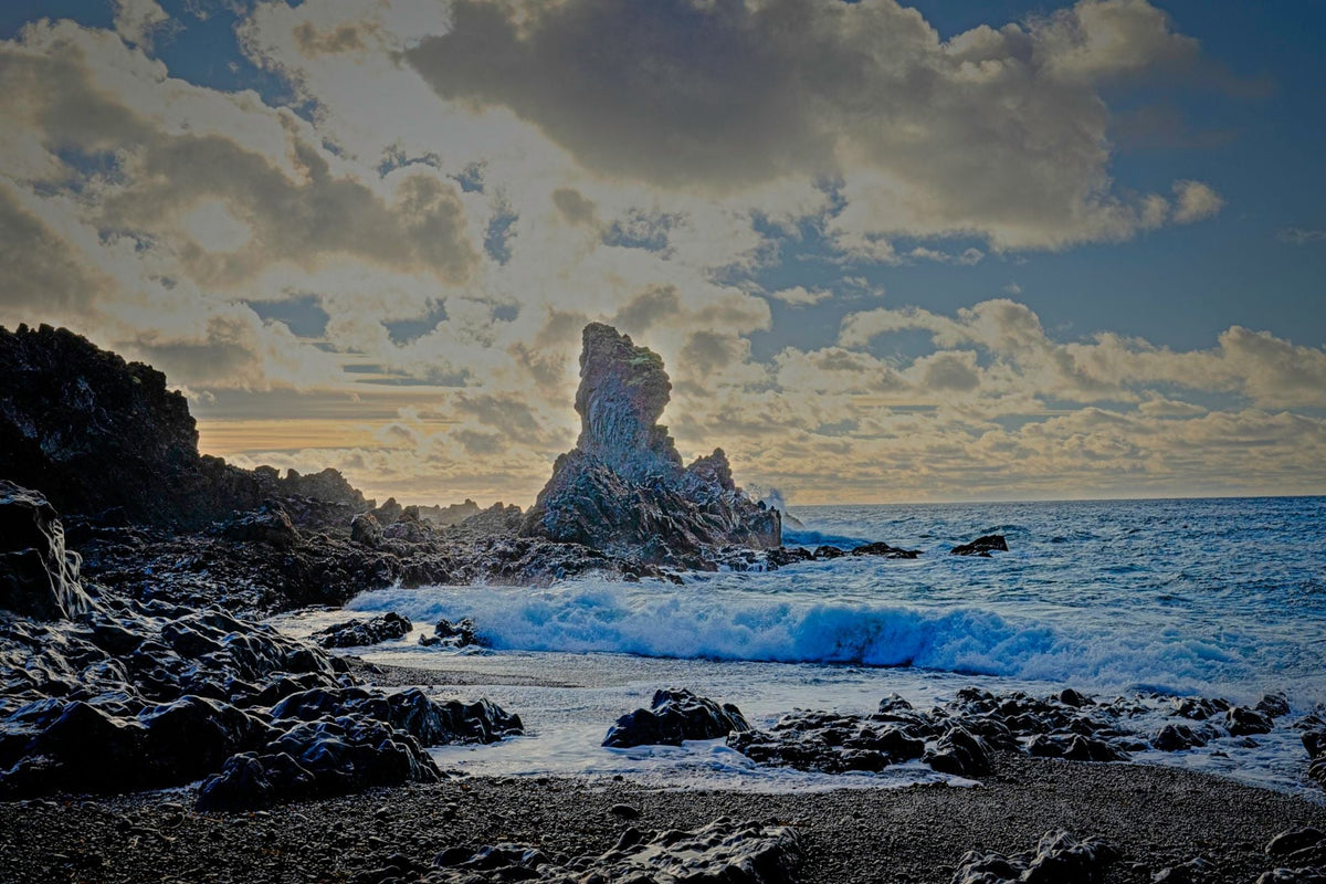 Rocks on a rocky beach with waves crashing against them under a cloudy sky.