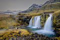 Kirkjufellsfoss waterfall cascading over a moss-covered basalt cliff, the striped slopes of Kirkjufell mountain rising behind under a pale sky