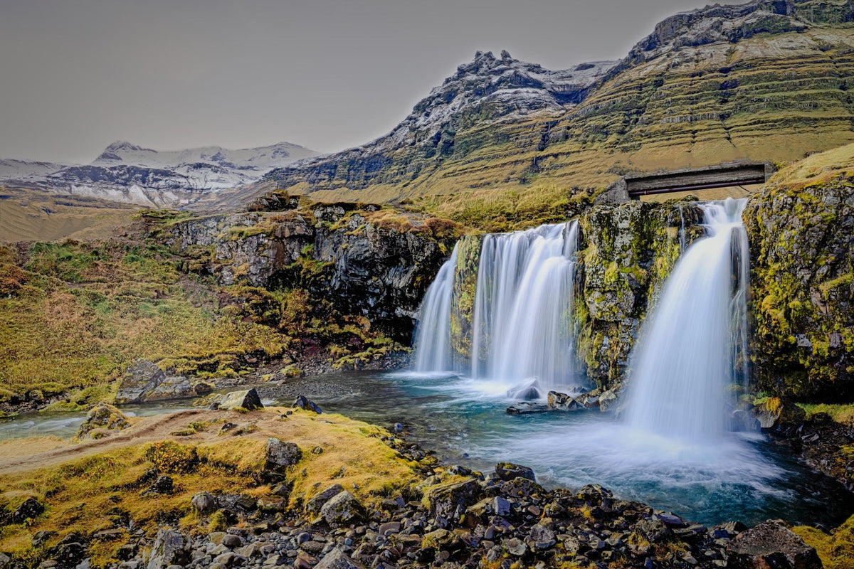 Kirkjufellsfoss waterfall cascading over a moss-covered basalt cliff, the striped slopes of Kirkjufell mountain rising behind under a pale sky