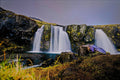 Twin waterfalls cascading over moss-covered basalt cliffs into a still pool at dusk, long exposure smoothing the falling water under a lavender Icelandic sky
