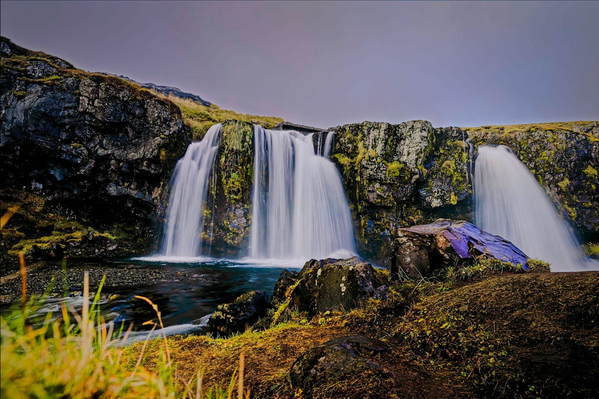 Twin waterfalls cascading over moss-covered basalt cliffs into a still pool at dusk, long exposure smoothing the falling water under a lavender Icelandic sky