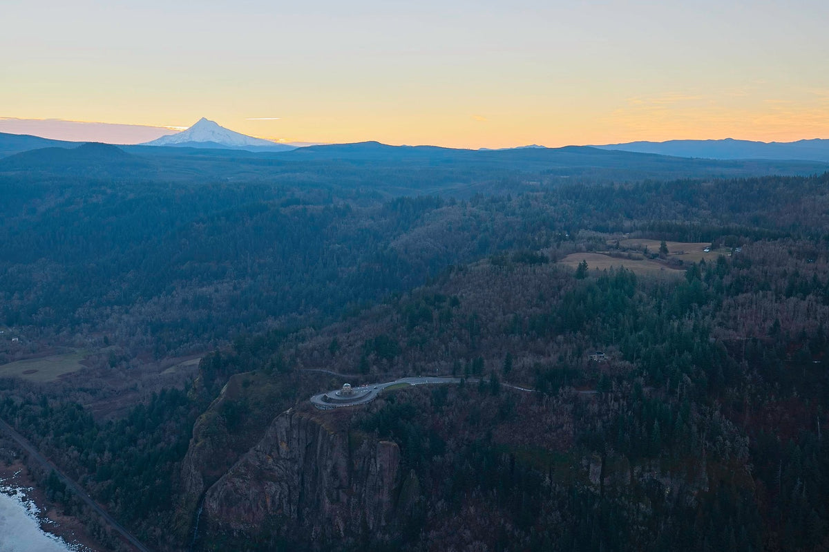 Mount Hood's distant peak catching last light beyond the forested Columbia River Gorge, a cliffside overlook road winding below at dusk