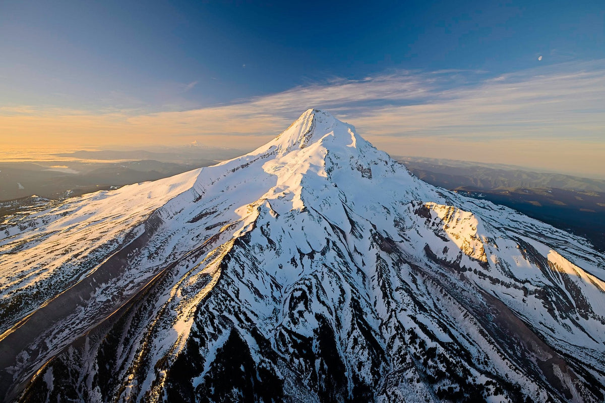 The full symmetric snow-covered cone of Mount Hood catching golden sunlight, dark forested ridges falling away toward the horizon