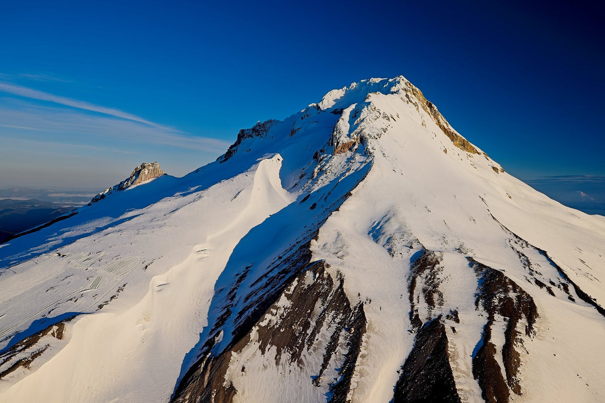 Golden light catching Mount Hood's summit crags beneath a deep sapphire sky, a rocky sub-peak visible to the west