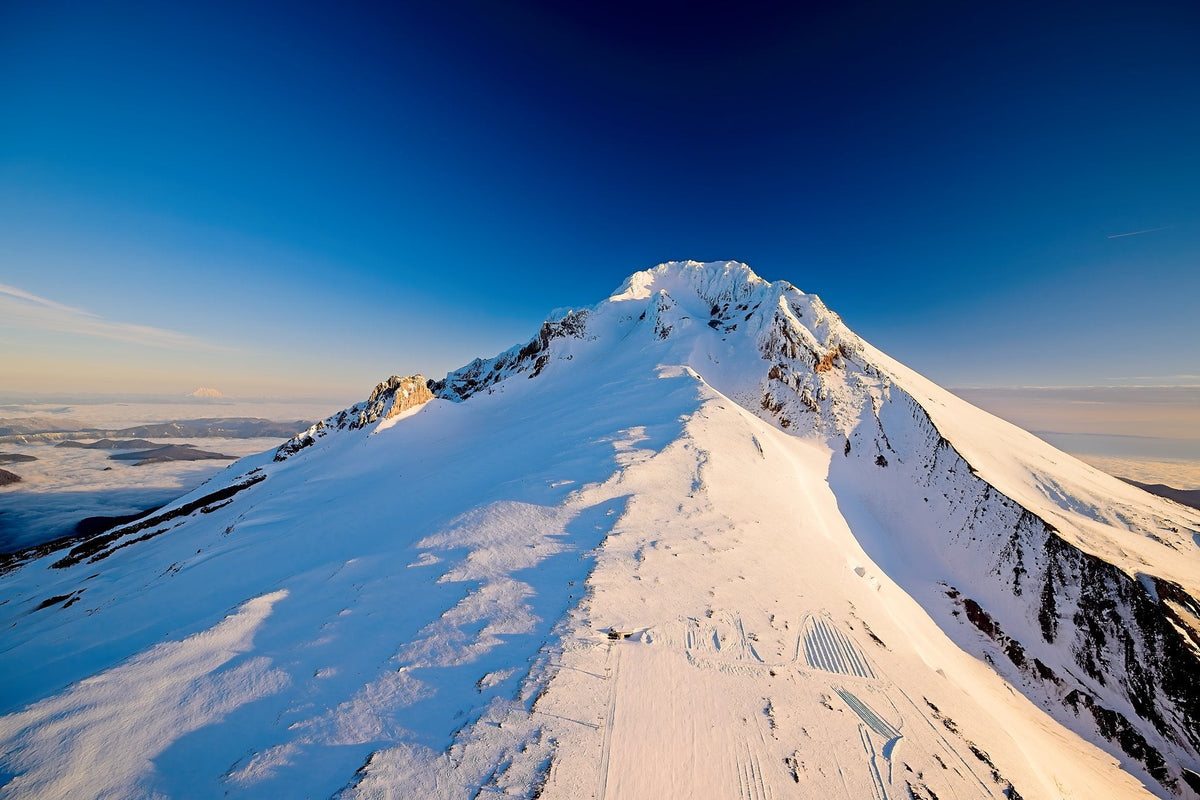Mount Hood's snow-covered summit crest under a deep blue midday sky, exposed rock along the ridge and ski infrastructure visible far below
