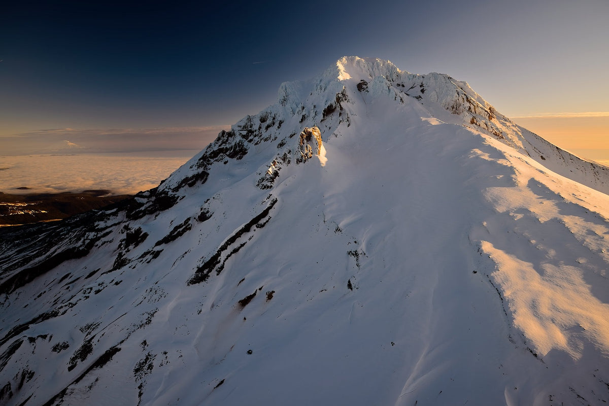 A broad, smooth snowfield flowing down from Mount Hood's summit, warm dawn light grazing one flank above a sea of clouds