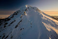 Dark volcanic rock exposed along Mount Hood's snow-covered summit ridge, a sea of clouds below and a warm glow along the horizon at dawn