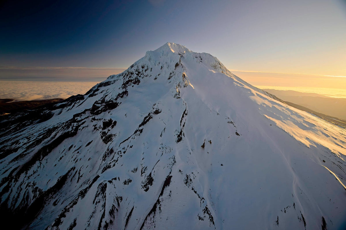 A ridge line of wind-carved snow running down from Mount Hood's summit at first light, layers of cloud and distant valley below