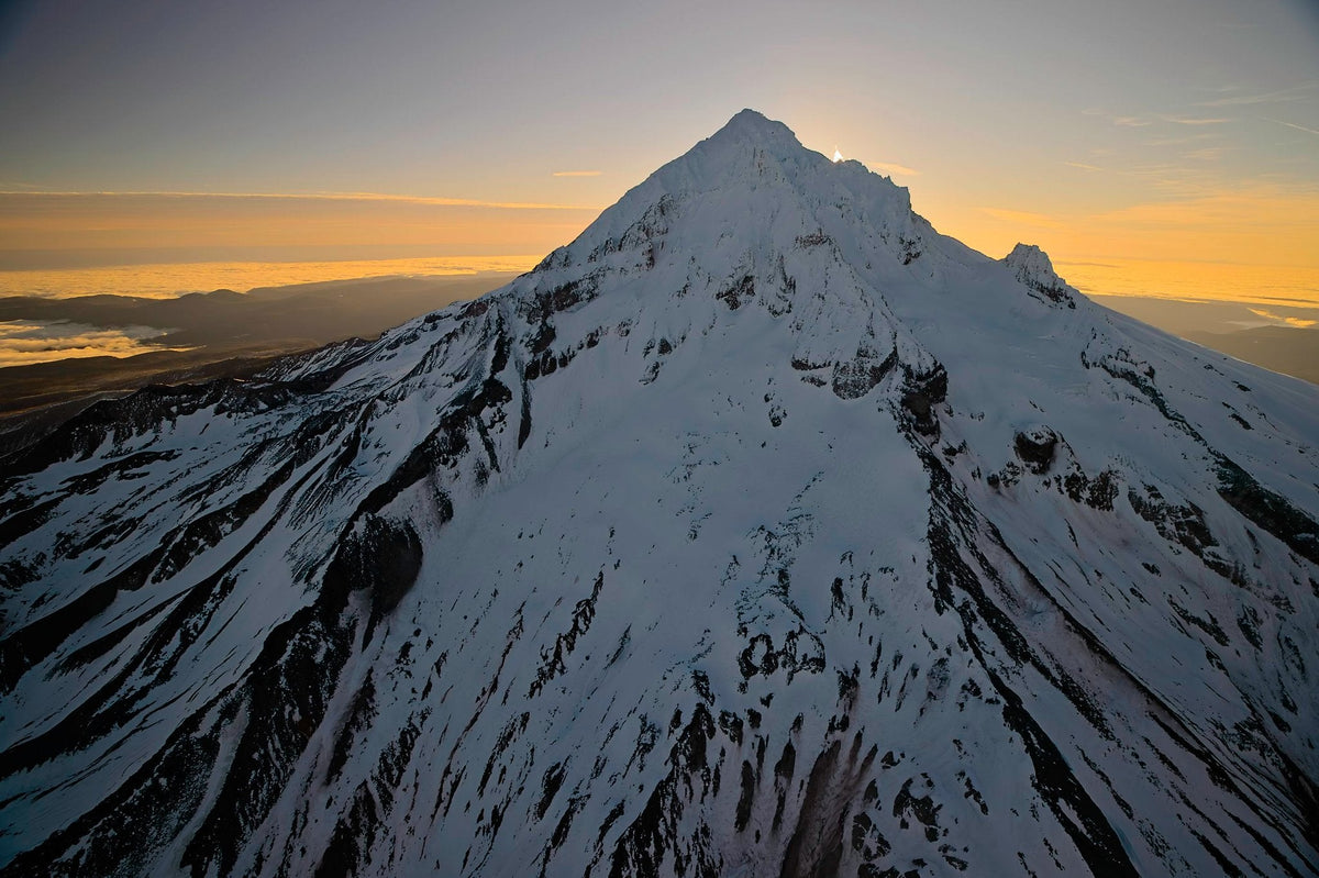 The sun breaking over Mount Hood's summit ridge in silhouette, amber light spreading along the horizon at sunrise