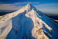 A sunlit rim of snow tracing Mount Hood's east face against a clear blue sky, a faint crescent moon near the summit