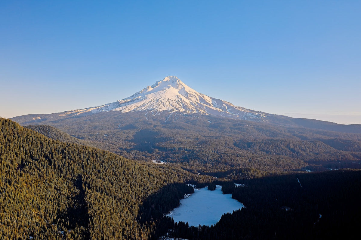 Mount Hood rising above dense evergreen forest, a frozen lake nestled in the valley below under a clear blue sky