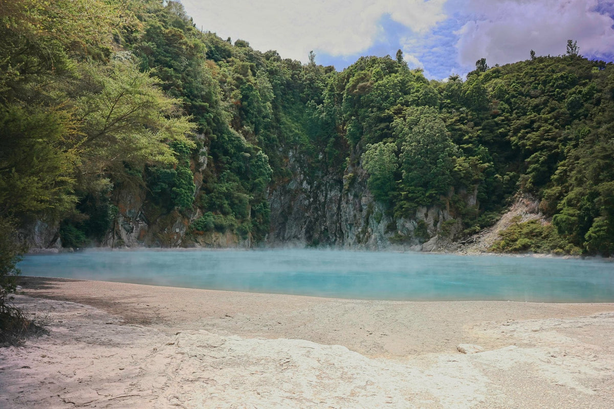 Steam rising off a vivid turquoise geothermal lake, pale mineral shoreline framed by forested cliffs