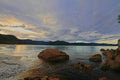 Rust-colored boulders anchored in the shallows of a still bay, layered coastal hills beneath a warm streaked sunset sky