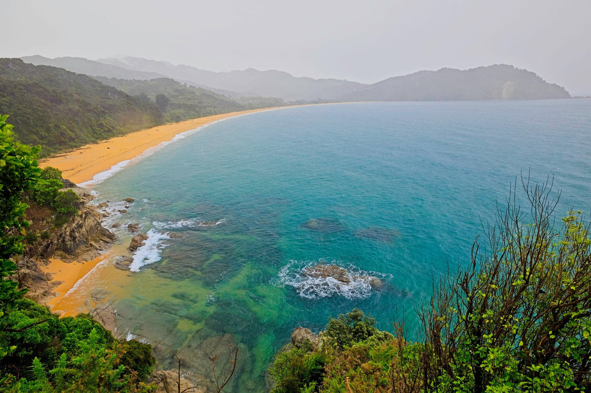 A golden sand beach curving along turquoise water, forested headlands fading into coastal haze