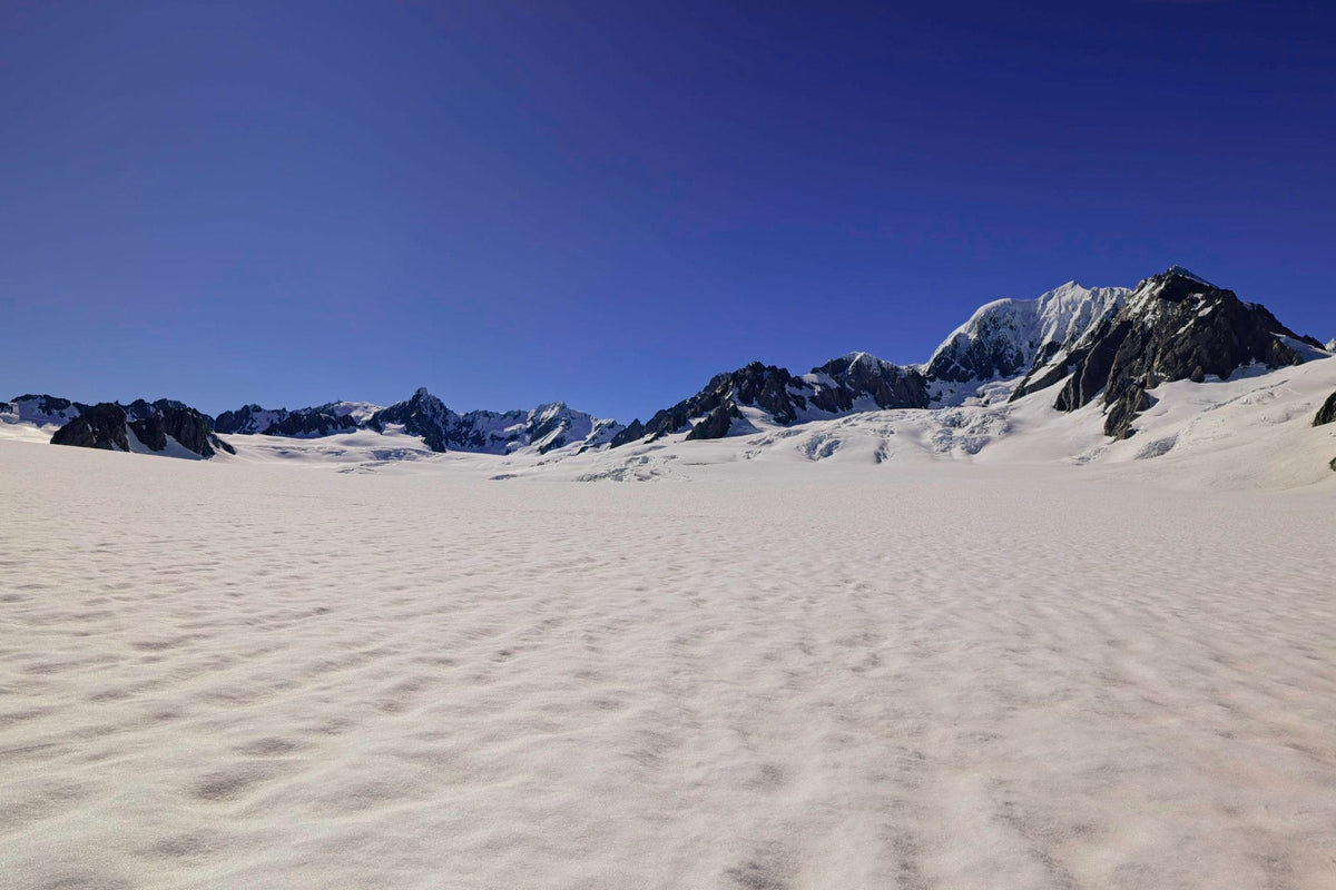 A vast glacier snowfield stretching beneath the jagged dark peaks of the Southern Alps under a deep blue sky