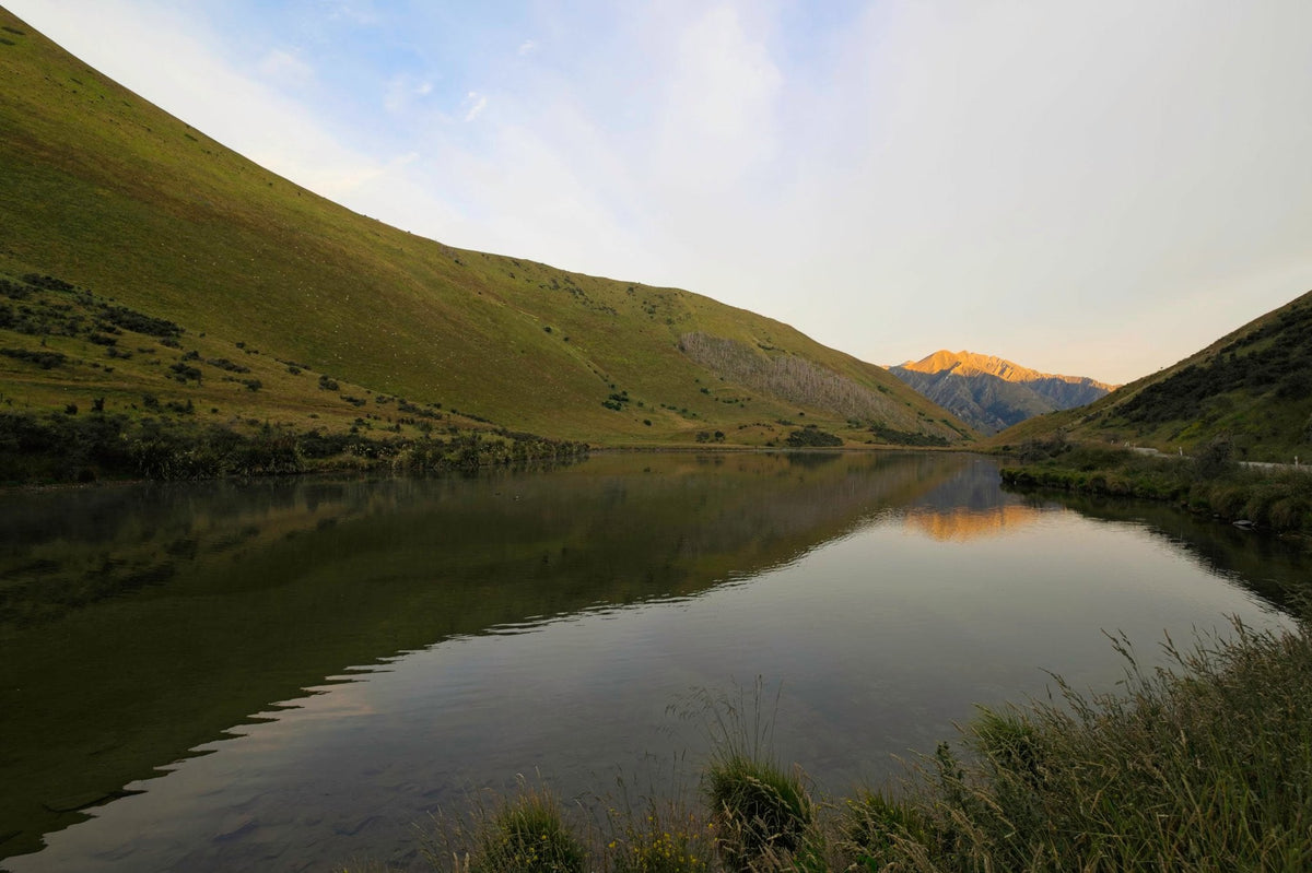 Grass-covered hills reflecting in the still water of Moke Lake, a distant peak catching the day's last sunlight
