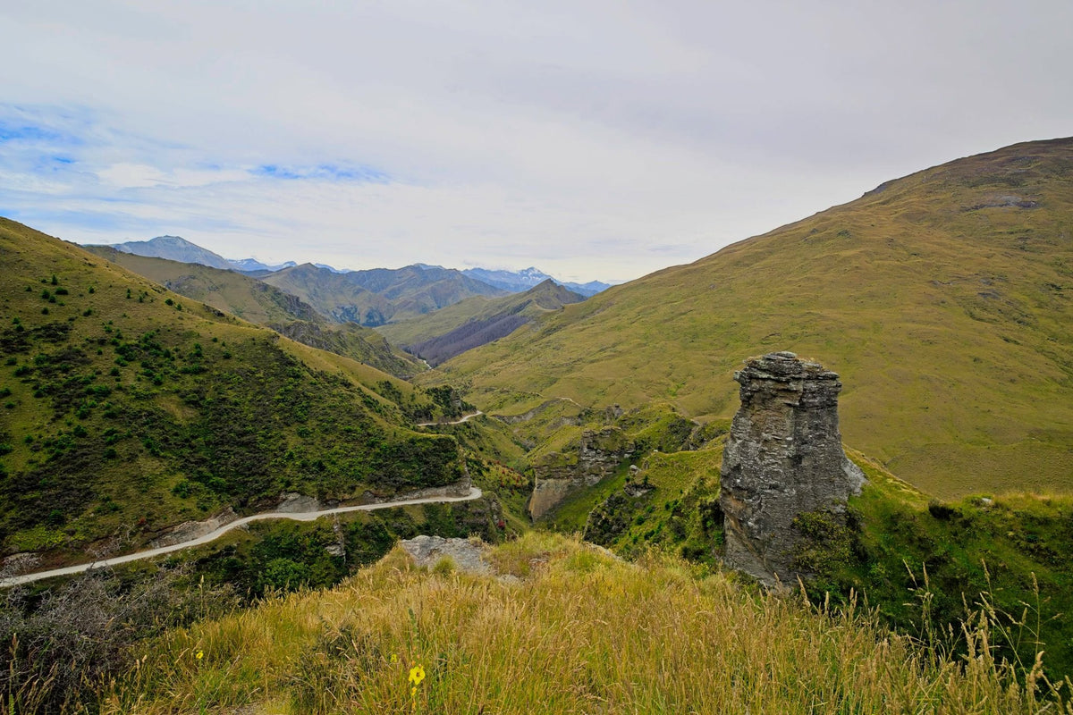 A narrow gravel road switchbacking down a steep golden hillside in Skippers Canyon, a rocky pinnacle in the foreground and layered ranges beyond