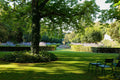 A formal Provençal garden with clipped box hedges, lavender borders and a stone fountain basin beneath a broad chestnut, green metal chairs on the lawn